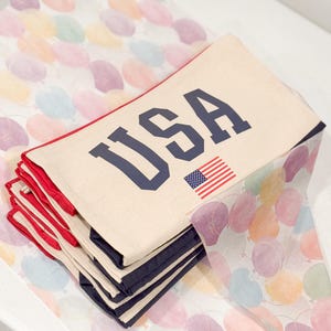 May include: A stack of cream-colored fabric items with the word "USA" in navy blue block letters and a small American flag. The items have red trim and navy blue borders. They are resting on a surface with a colorful balloon pattern.