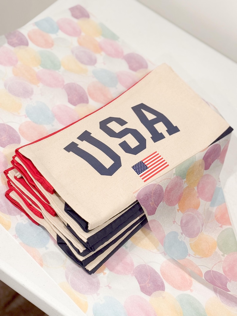 May include: A stack of cream-colored fabric items with the word "USA" in navy blue block letters and a small American flag. The items have red trim and navy blue borders. They are resting on a surface with a colorful balloon pattern.
