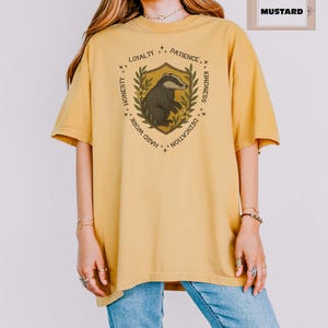 Puede incluir: Camiseta de color mostaza con un dise&ntilde;o de escudo y una ilustraci&oacute;n de tej&oacute;n. El escudo incluye el texto "Lealtad, Paciencia, Amabilidad, Dedicaci&oacute;n, Trabajo duro, Honestidad". La camiseta es de corte holgado y tiene mangas cortas.