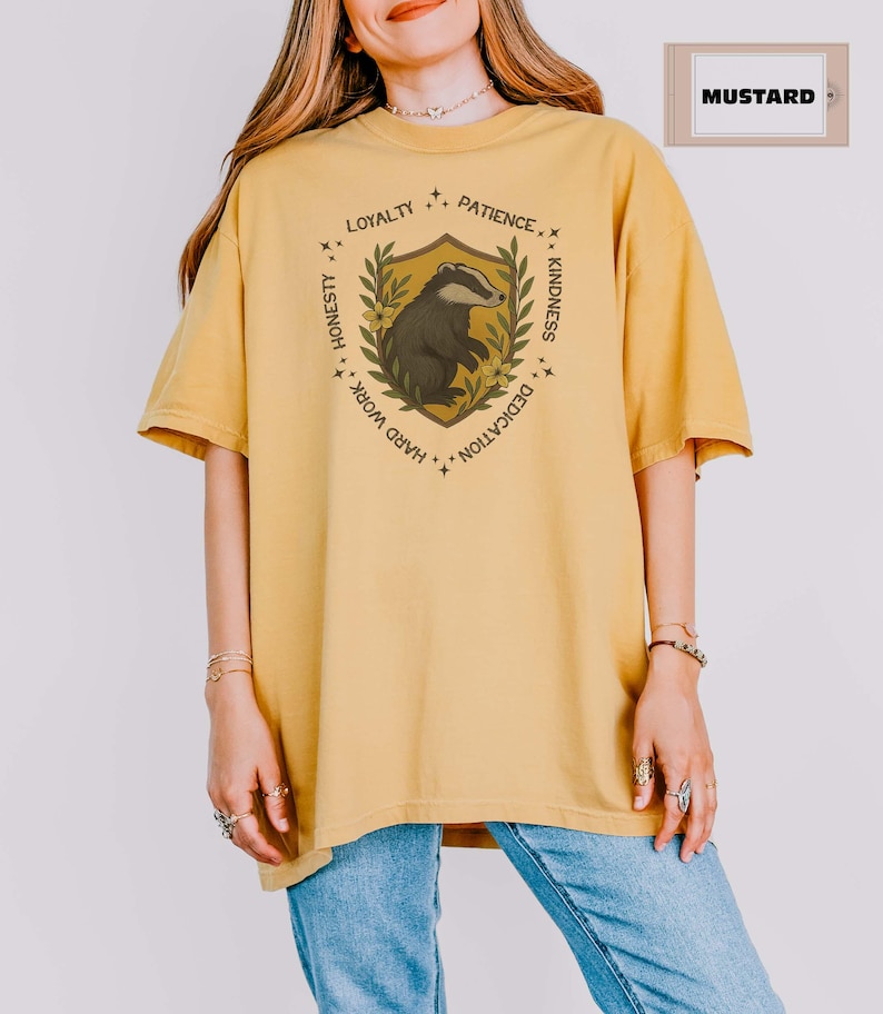 Puede incluir: Camiseta de color mostaza con un dise&ntilde;o de escudo y una ilustraci&oacute;n de tej&oacute;n. El escudo incluye el texto "Lealtad, Paciencia, Amabilidad, Dedicaci&oacute;n, Trabajo duro, Honestidad". La camiseta es de corte holgado y tiene mangas cortas.