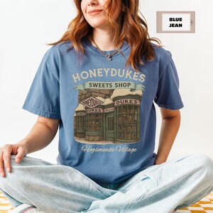 Op de afbeelding: Een blauw T-shirt met een afbeelding van Honeydukes Sweets Shop uit de Harry Potter-serie. De afbeelding bevat de tekst "Honeydukes Sweets Shop" en "Hogsmeade Village".