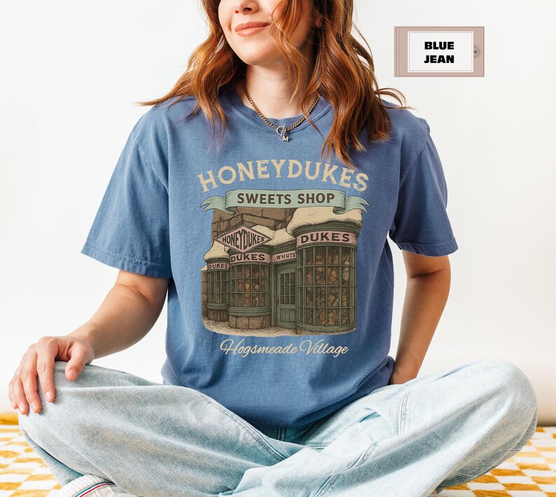 Op de afbeelding: Een blauw T-shirt met een afbeelding van Honeydukes Sweets Shop uit de Harry Potter-serie. De afbeelding bevat de tekst "Honeydukes Sweets Shop" en "Hogsmeade Village".