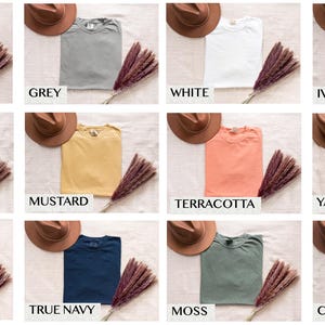 Op de afbeelding: Een verzameling van tien t-shirts in verschillende kleuren, waaronder peper, grijs, wit, ivoor, boter, mosterd, terracotta, yam, spijkerblauw, donkerblauw, mos en karmijnrood. De t-shirts liggen op een lichtbruine ondergrond met gedroogde bloemen.