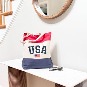 May include: A canvas tote bag with a red and navy blue color block design. The bag features the word "USA" in navy blue block letters, with a small American flag graphic. Sunglasses rest on a white surface.