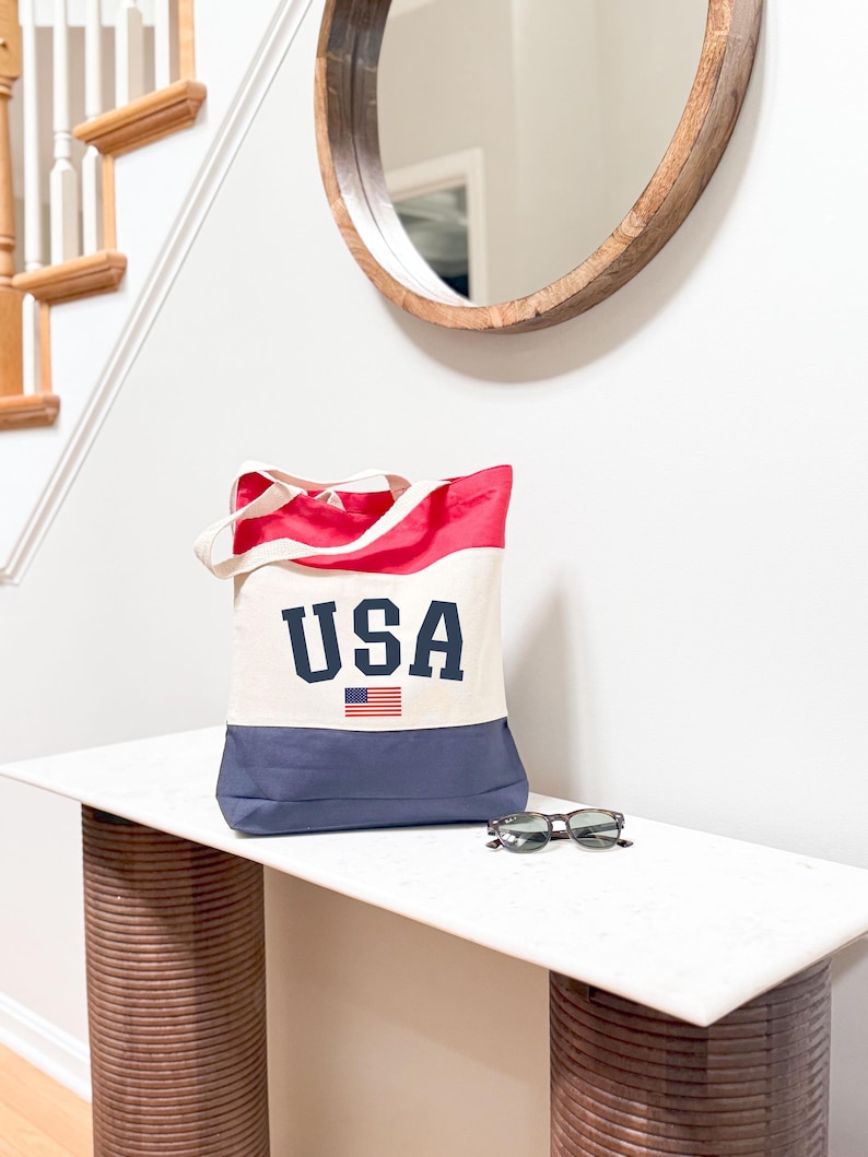 May include: A canvas tote bag with a red and navy blue color block design. The bag features the word "USA" in navy blue block letters, with a small American flag graphic. Sunglasses rest on a white surface.