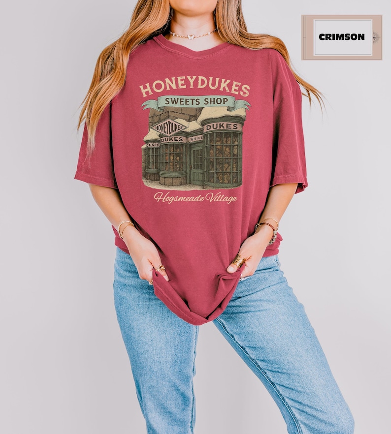 May include: A crimson t-shirt with a graphic of Honeydukes Sweets Shop from Hogsmeade Village. The graphic is a vintage-style illustration of the shop's storefront with the text 'Honeydukes Sweets Shop' and 'Hogsmeade Village'.