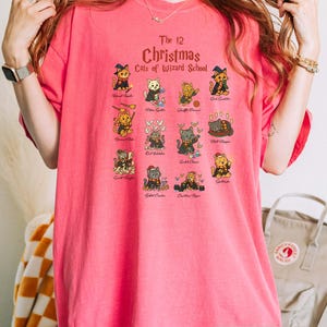 Woman wearing a water melon t-shirt featuring a holiday design titled “The 12 Christmas Cats of Wizard School,” showing cute illustrated cats dressed as magical characters. Festive Christmas decor in the background including tree, pillows, and seasonal accents.