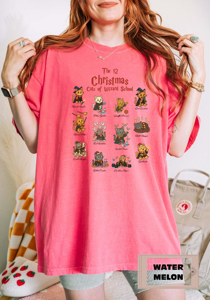Woman wearing a water melon t-shirt featuring a holiday design titled “The 12 Christmas Cats of Wizard School,” showing cute illustrated cats dressed as magical characters. Festive Christmas decor in the background including tree, pillows, and seasonal accents.