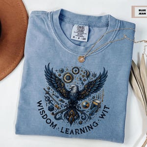 May include: A blue jean t-shirt with a graphic of a stylized eagle surrounded by various symbols and the text "Wisdom: Learning: Wit".