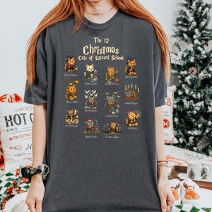 Woman wearing a pepper t-shirt featuring a holiday design titled “The 12 Christmas Cats of Wizard School,” showing cute illustrated cats dressed as magical characters. Festive Christmas decor in the background including tree, pillows, and seasonal accents.