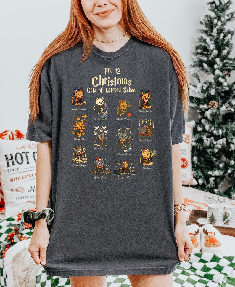 Woman wearing a pepper t-shirt featuring a holiday design titled “The 12 Christmas Cats of Wizard School,” showing cute illustrated cats dressed as magical characters. Festive Christmas decor in the background including tree, pillows, and seasonal accents.