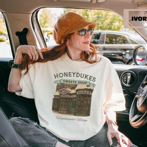 Op de afbeelding: Een wit T-shirt met een afbeelding van Honeydukes Sweets Shop uit de Harry Potter-serie. De afbeelding toont de gevel van de winkel met de tekst "Honeydukes Sweets Shop" en "Hogsmeade Village".