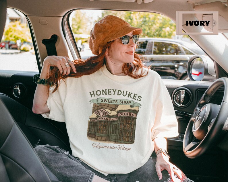 Op de afbeelding: Een wit T-shirt met een afbeelding van Honeydukes Sweets Shop uit de Harry Potter-serie. De afbeelding toont de gevel van de winkel met de tekst "Honeydukes Sweets Shop" en "Hogsmeade Village".