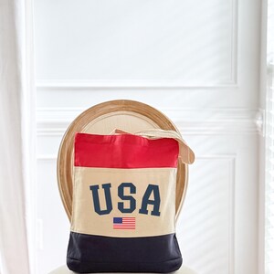 May include: A canvas tote bag with a patriotic design. The top section is red, the middle is beige, and the bottom is navy blue. The word "USA" is printed in navy blue block letters, with a small American flag below.
