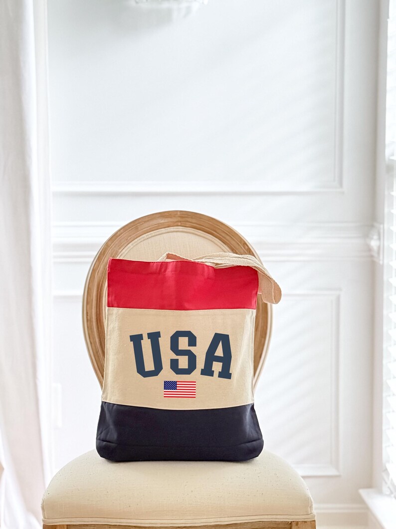 May include: A canvas tote bag with a patriotic design. The top section is red, the middle is beige, and the bottom is navy blue. The word "USA" is printed in navy blue block letters, with a small American flag below.