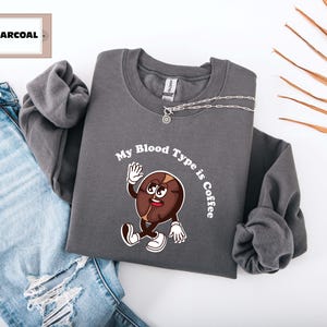 Gray sweatshirt featuring a playful design with a smiling cartoon coffee bean character waving and walking. Above the illustration, bold curved text reads 'My Blood Type is Coffee' in dark brown.