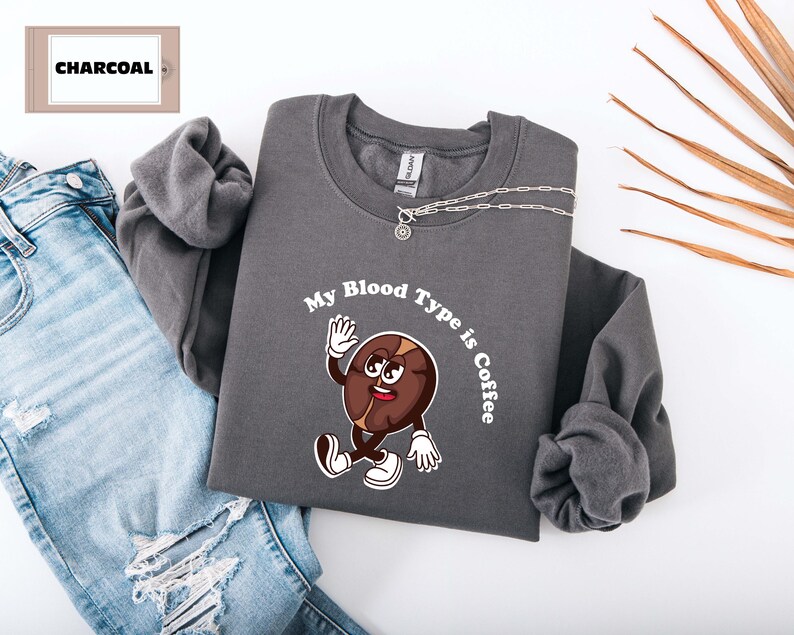 Gray sweatshirt featuring a playful design with a smiling cartoon coffee bean character waving and walking. Above the illustration, bold curved text reads 'My Blood Type is Coffee' in dark brown.
