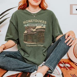 Op de afbeelding: Een groen T-shirt met een afbeelding van Honeydukes Sweetshop uit de Harry Potter-serie. De afbeelding toont een gebouw met de tekst "Honeydukes Sweetshop" en "Hogsmeade Village".