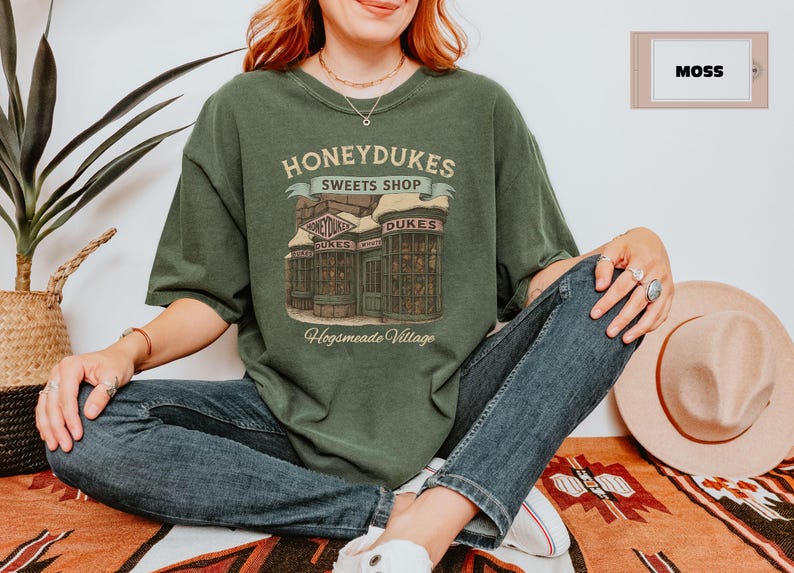 Op de afbeelding: Een groen T-shirt met een afbeelding van Honeydukes Sweetshop uit de Harry Potter-serie. De afbeelding toont een gebouw met de tekst "Honeydukes Sweetshop" en "Hogsmeade Village".
