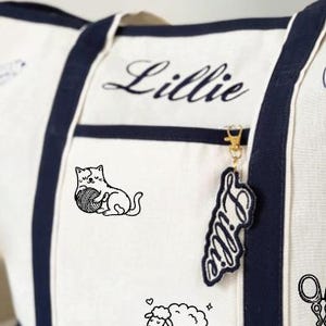 May include: A cream-coloured canvas tote bag with navy blue trim and handles. The bag features the name Lillie embroidered in navy blue script, a black line drawing of a cat with a ball of yarn, a sheep, and a pair of scissors. A matching name tag hangs from the zip.