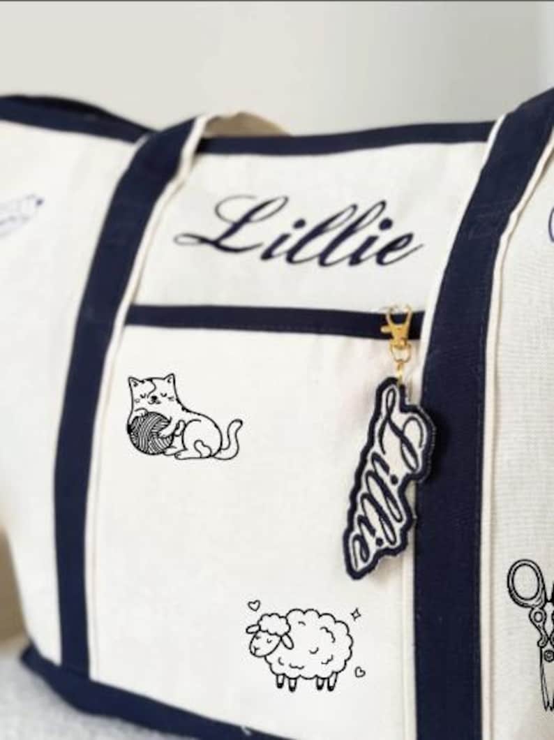 May include: A cream-coloured canvas tote bag with navy blue trim and handles. The bag features the name Lillie embroidered in navy blue script, a black line drawing of a cat with a ball of yarn, a sheep, and a pair of scissors. A matching name tag hangs from the zip.