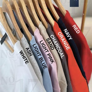 以下が含まれることがあります： 木製のハンガーに掛けられた10枚のTシャツのセット。Tシャツは、白、アッシュ、ライトブルー、ライトピンク、サンド、チャコール、オレンジ、ネイビー、レッドなど、さまざまな色があります。Tシャツには色が表示されています。