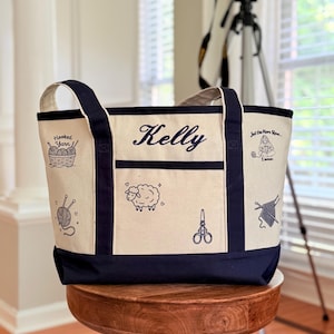May include: A cream-coloured canvas tote bag with navy blue trim and handles. The bag features the name Kelly embroidered in navy blue, along with black line art illustrations of yarn, a sheep, scissors, and text reading Hooked Yarn and Just One More Row I swear.