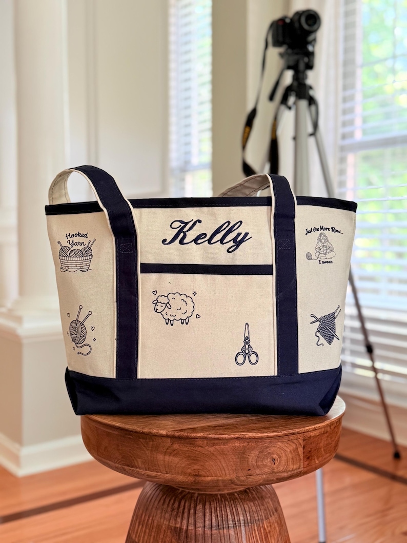 May include: A cream-coloured canvas tote bag with navy blue trim and handles. The bag features the name Kelly embroidered in navy blue, along with black line art illustrations of yarn, a sheep, scissors, and text reading Hooked Yarn and Just One More Row I swear.