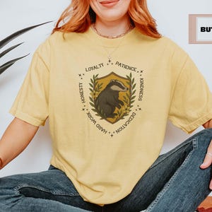 Puede incluir: Camiseta amarilla clara con un dise&ntilde;o de escudo que presenta un tej&oacute;n, rodeado de texto que incluye "Lealtad", "Paciencia" y "Amabilidad". La camiseta es de color amarillo claro y tiene un ajuste relajado.