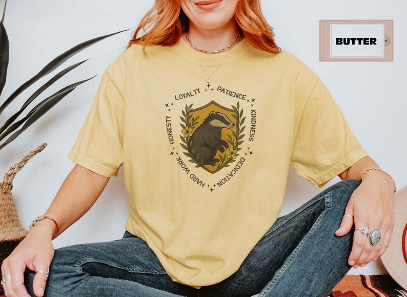 Puede incluir: Camiseta amarilla clara con un dise&ntilde;o de escudo que presenta un tej&oacute;n, rodeado de texto que incluye "Lealtad", "Paciencia" y "Amabilidad". La camiseta es de color amarillo claro y tiene un ajuste relajado.