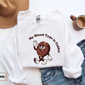 White sweatshirt featuring a playful design with a smiling cartoon coffee bean character waving and walking. Above the illustration, bold curved text reads 'My Blood Type is Coffee' in dark brown.