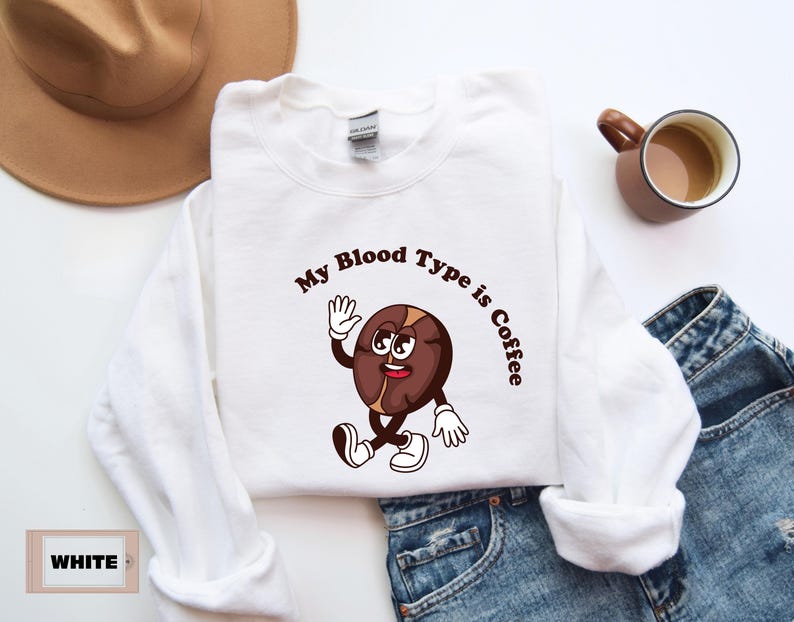 White sweatshirt featuring a playful design with a smiling cartoon coffee bean character waving and walking. Above the illustration, bold curved text reads 'My Blood Type is Coffee' in dark brown.