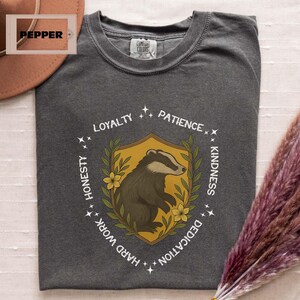 Puede incluir: Camiseta gris oscuro con un emblema de tej&oacute;n. El emblema es un tej&oacute;n dentro de un escudo, rodeado por las palabras "Lealtad, Paciencia, Amabilidad, Dedicaci&oacute;n, Trabajo duro, Honestidad". El escudo es amarillo con detalles florales verdes y amarillos.