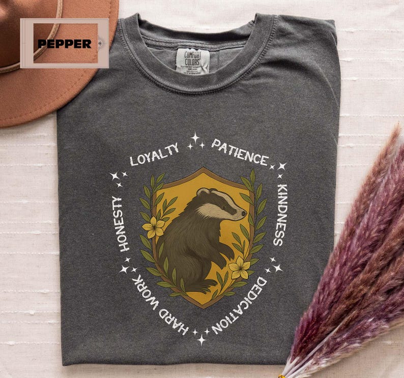 Puede incluir: Camiseta gris oscuro con un emblema de tej&oacute;n. El emblema es un tej&oacute;n dentro de un escudo, rodeado por las palabras "Lealtad, Paciencia, Amabilidad, Dedicaci&oacute;n, Trabajo duro, Honestidad". El escudo es amarillo con detalles florales verdes y amarillos.