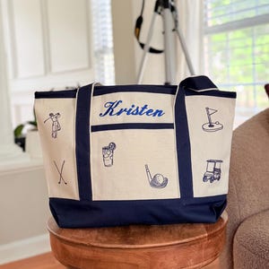 May include: A canvas tote bag with a navy blue base and trim, featuring embroidered golf-themed illustrations and the name "Kristen" in blue script. The bag includes images of golf clubs, a golf cart, and a golf ball.