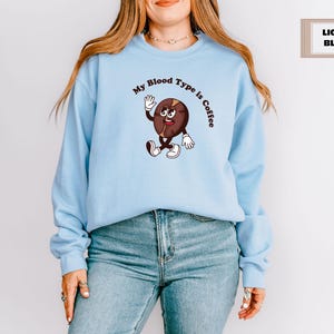 Navy sweatshirt featuring a playful design with a smiling cartoon coffee bean character waving and walking. Above the illustration, bold curved text reads 'My Blood Type is Coffee' in dark brown.