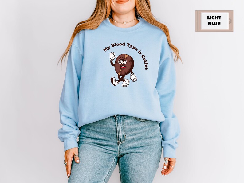 Navy sweatshirt featuring a playful design with a smiling cartoon coffee bean character waving and walking. Above the illustration, bold curved text reads 'My Blood Type is Coffee' in dark brown.