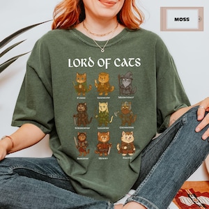 May include: Moss green t-shirt with the text "LORD OF CATS" above illustrated cat characters. Each cat is dressed in a costume with a name below. The shirt is a relaxed fit.