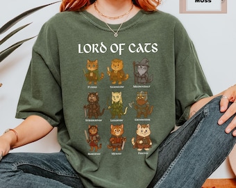 May include: Moss green t-shirt with the text "LORD OF CATS" above illustrated cat characters. Each cat is dressed in a costume with a name below. The shirt is a relaxed fit.