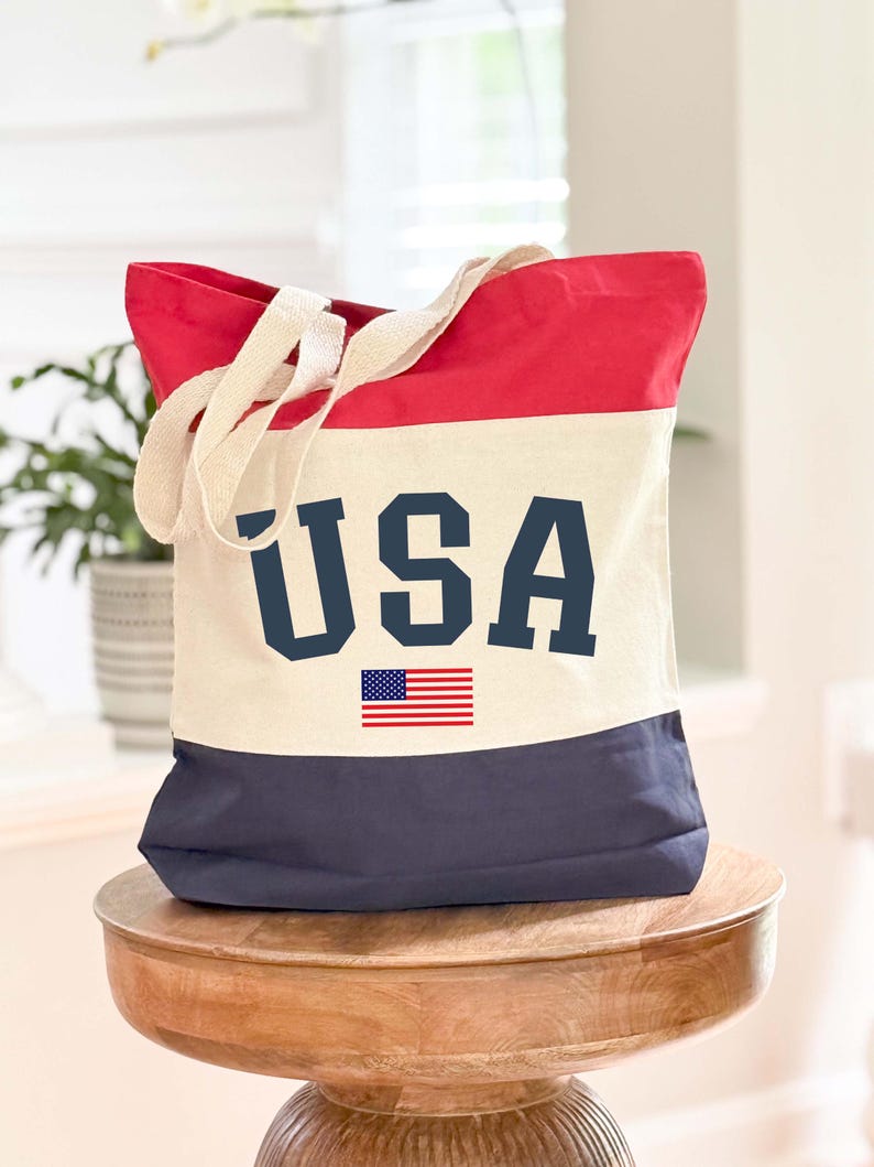 May include: A red, white, and blue tote bag with the word "USA" in navy blue block letters and a small American flag graphic. The bag has cream-colored handles and is sitting on a wooden stool.