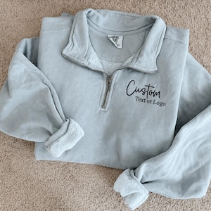 May include: A light blue quarter-zip sweatshirt with a folded collar and long sleeves. The garment features a dark blue embroidered script that reads Custom and the text Text or Logo below it on the upper left chest area near the zipper.