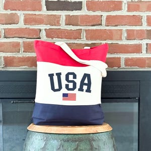May include: A patriotic tote bag with a red top, white center, and navy blue bottom. The word "USA" is printed in navy blue block letters, with a small American flag beneath it. The bag has a white handle.