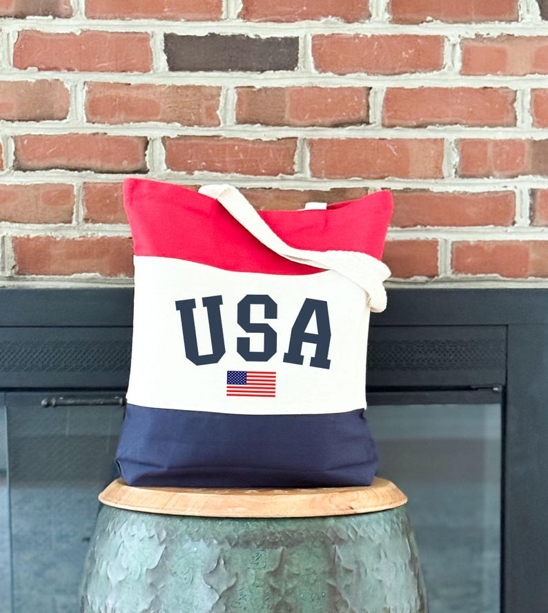 May include: A patriotic tote bag with a red top, white center, and navy blue bottom. The word "USA" is printed in navy blue block letters, with a small American flag beneath it. The bag has a white handle.