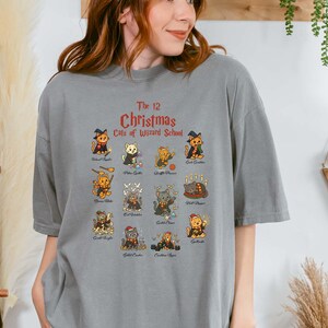 Woman wearing a gray t-shirt featuring a holiday design titled “The 12 Christmas Cats of Wizard School,” showing cute illustrated cats dressed as magical characters. Festive Christmas decor in the background including tree, pillows, and seasonal accents.
