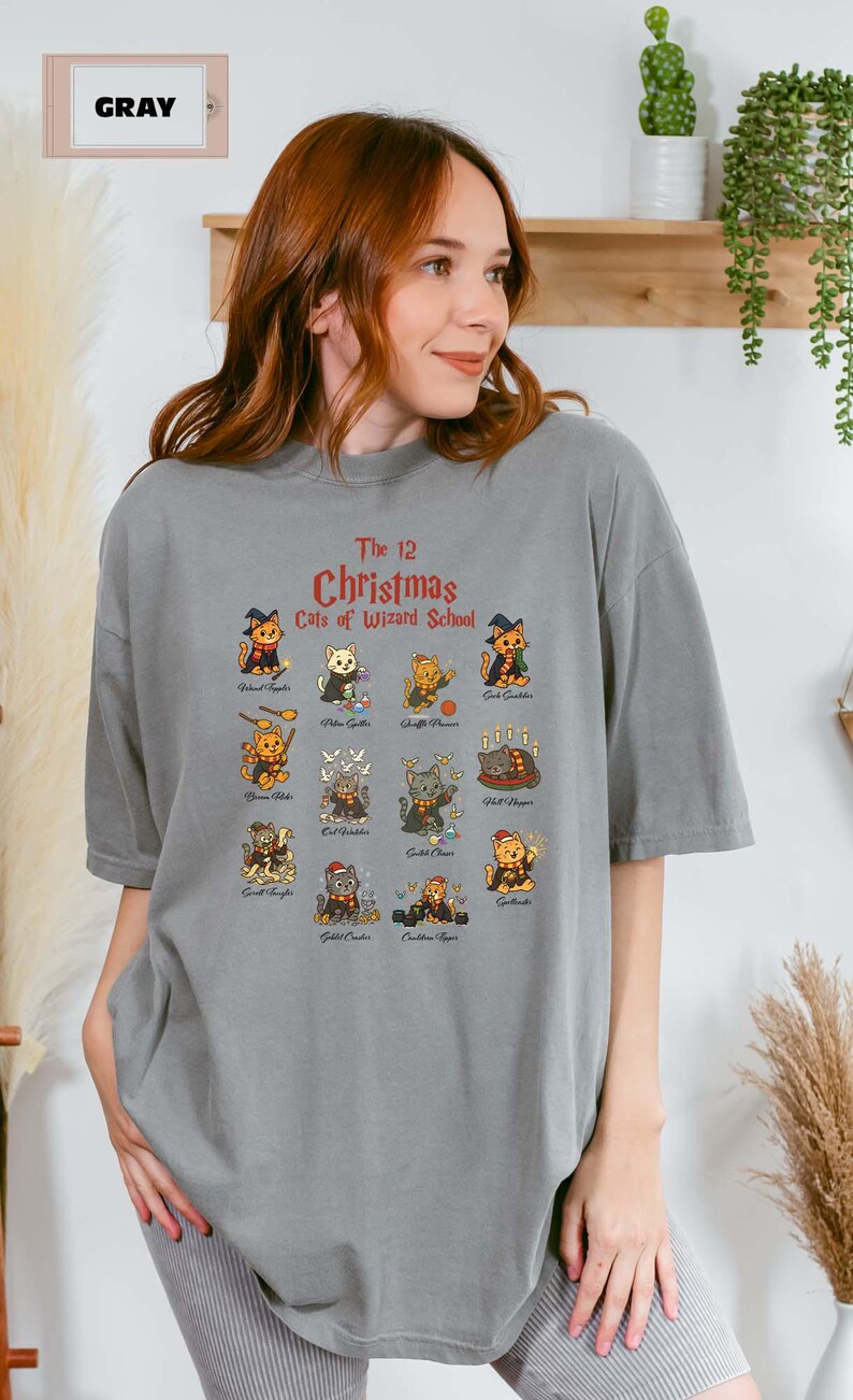 Woman wearing a gray t-shirt featuring a holiday design titled “The 12 Christmas Cats of Wizard School,” showing cute illustrated cats dressed as magical characters. Festive Christmas decor in the background including tree, pillows, and seasonal accents.