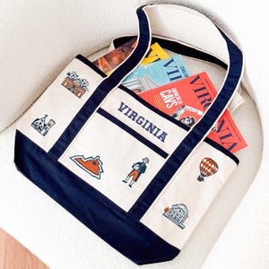May include: A canvas tote bag with a cream-colored body and navy blue trim. The bag features the word "VIRGINIA" in navy blue block letters and various state-themed illustrations. Several magazines are visible inside the bag.