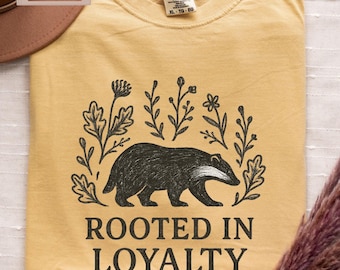 Puede incluir: Una camiseta amarilla mostaza con una ilustración de tejón negro rodeada de flores y hojas. El texto "Rooted in Loyalty" está impreso debajo del tejón.