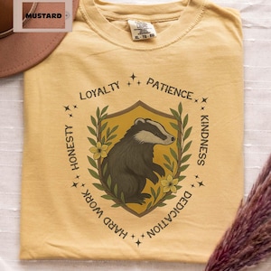 Puede incluir: Camiseta de color mostaza con una ilustraci&oacute;n de tej&oacute;n dentro de un dise&ntilde;o de escudo. El escudo est&aacute; rodeado por las palabras "Lealtad, Paciencia, Amabilidad, Dedicaci&oacute;n, Trabajo duro, Honestidad". La camiseta es talla XL-TG-EG.