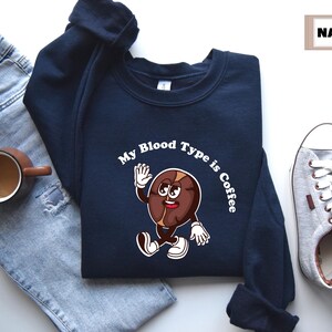 Navy sweatshirt featuring a playful design with a smiling cartoon coffee bean character waving and walking. Above the illustration, bold curved text reads 'My Blood Type is Coffee' in dark brown.