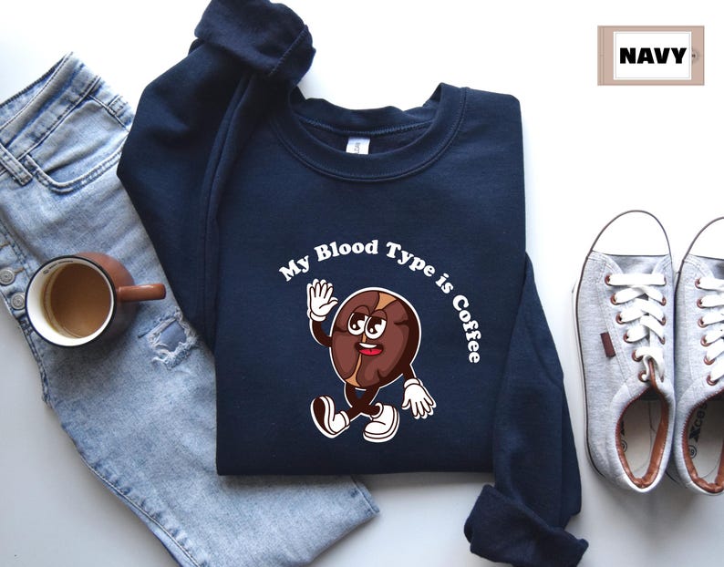 Navy sweatshirt featuring a playful design with a smiling cartoon coffee bean character waving and walking. Above the illustration, bold curved text reads 'My Blood Type is Coffee' in dark brown.